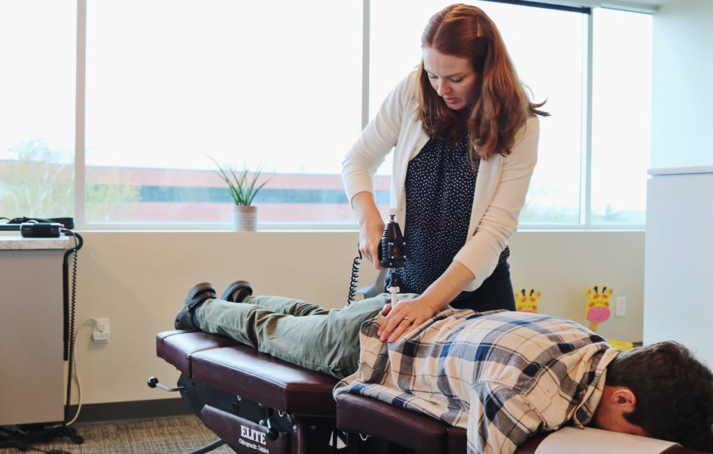 Chiropractor using an adjusting instrument on a patient’s upper back while lying face down.