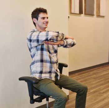 Patient seated in office chair performing upper-body mobility stretch with arms crossed.