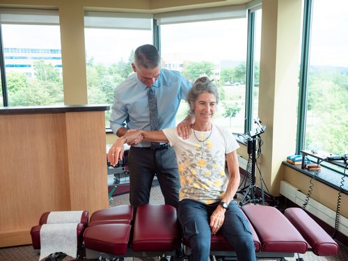 Chiropractor performs a shoulder adjustment on a seated patient beside a treatment table.